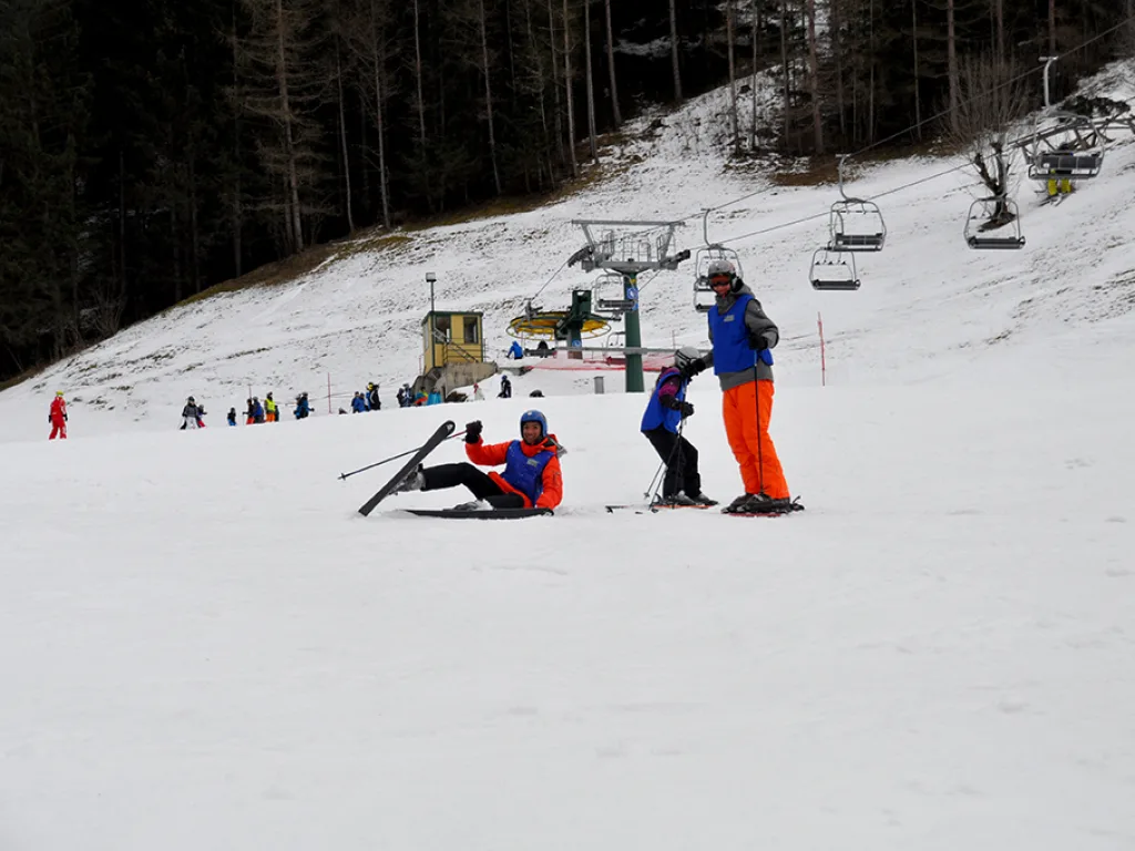 Zwei Personen helfen einer gestürzten Person auf der Piste.